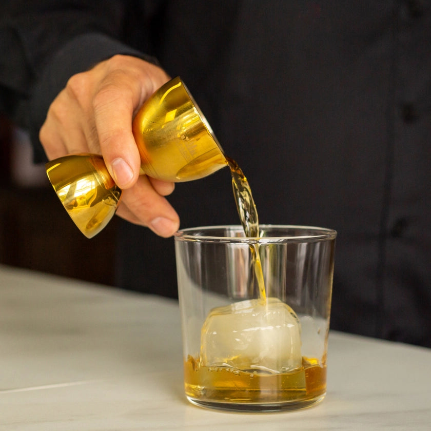 Hand pouring a golden liquid from a small container into a glass with ice cubes on a neutral background