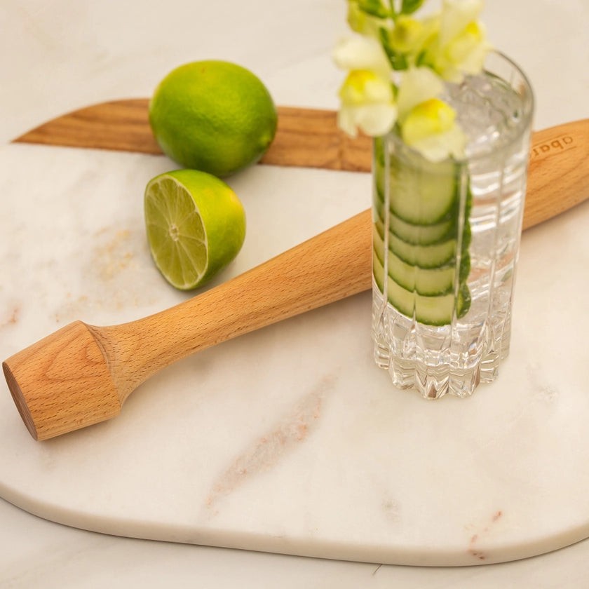 Wooden muddler, limes, and a glass with cucumber slices on a marble surface