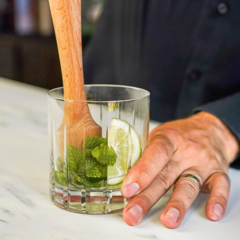 Glass with lime and mint being stirred with a wooden spoon, held by a person.