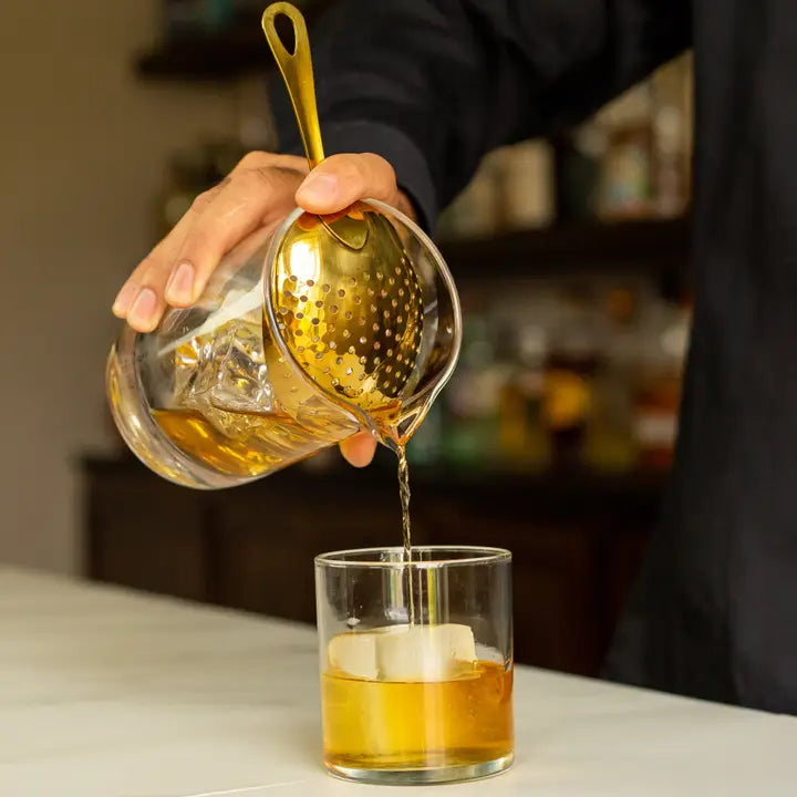 Person pouring liquid from a glass container with a strainer into a glass on a countertop.