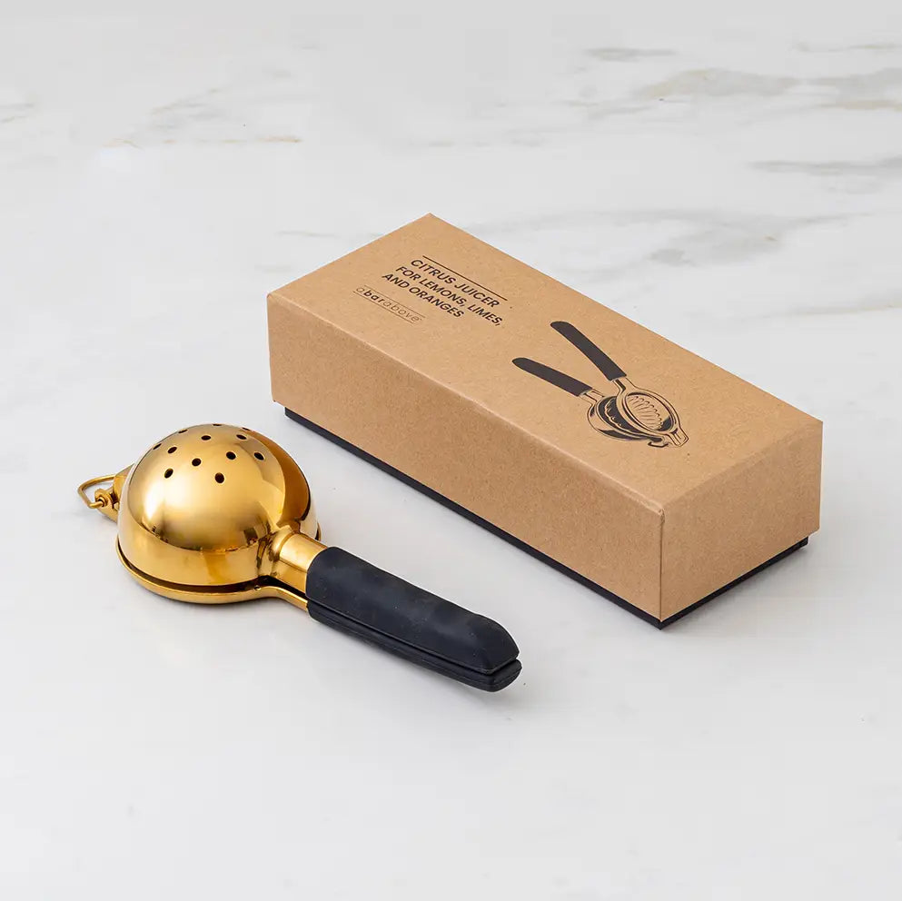 Gold citrus juicer with black handle next to its cardboard box on a light gray background