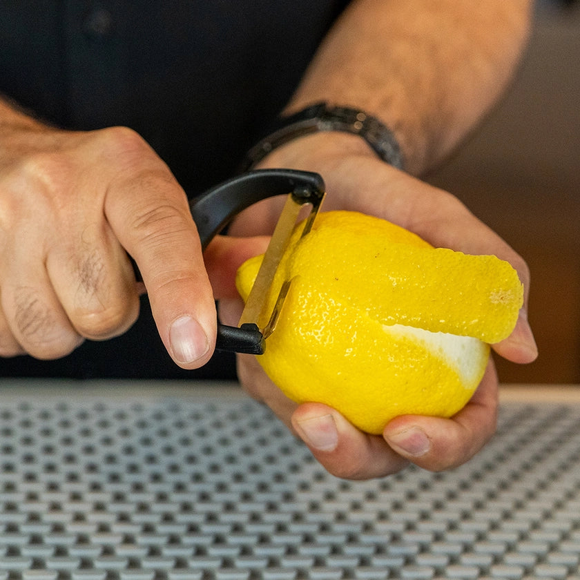 Person using a peeler to remove skin from a lemon on a textured surface.