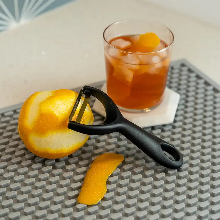 Orange being peeled with a peeler next to a glass of iced tea on a textured surface.