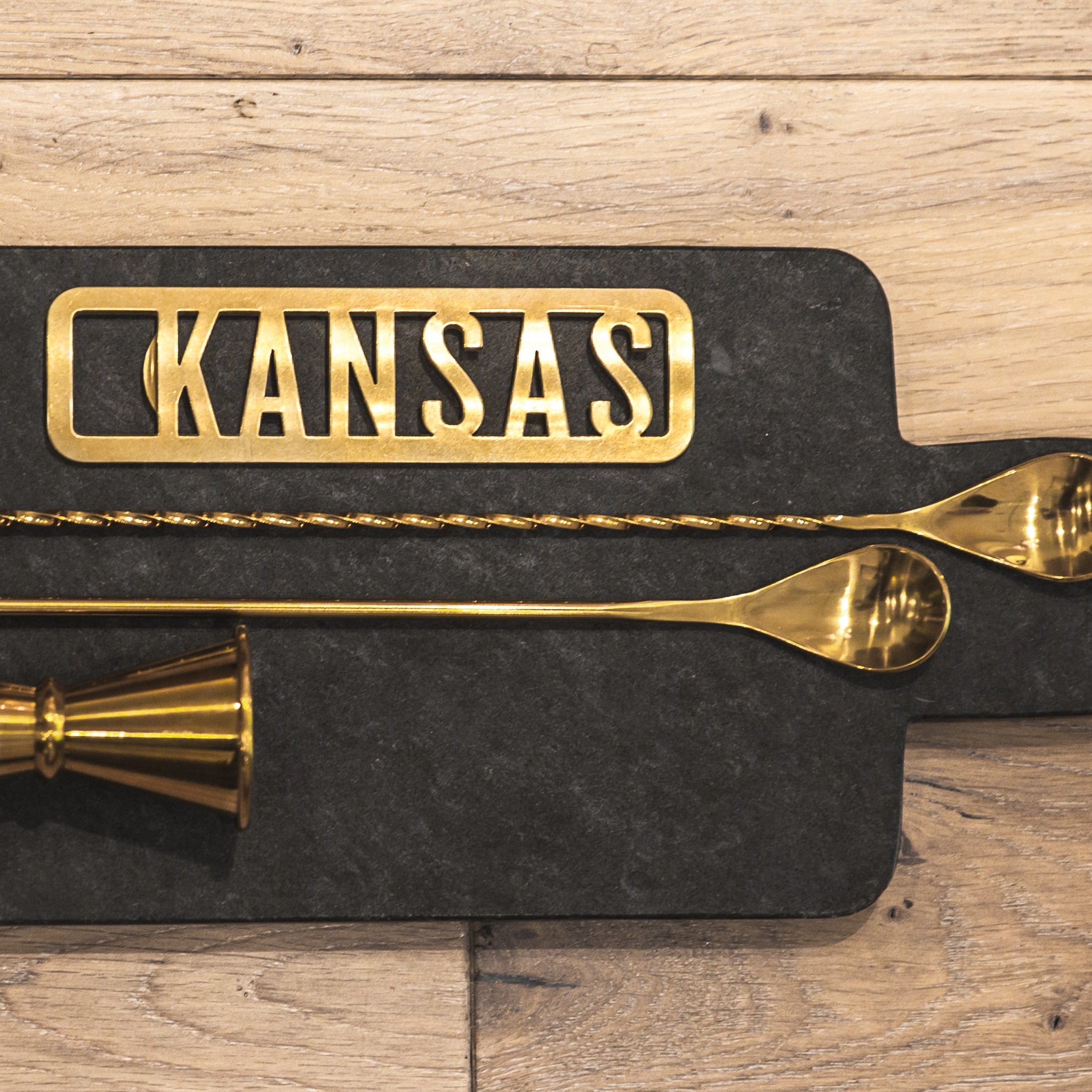 Gold kitchen utensils on a black mat with 'KANSAS' engraving on a wooden surface